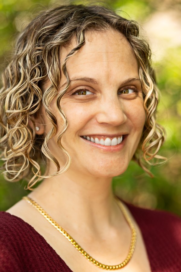 Meredith smiling in a deep red top, curly highlighted hair framed by soft garden greens