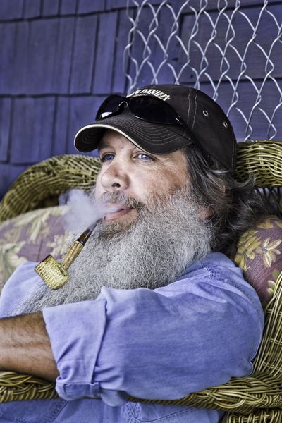 A bearded man with bright blue eyes smoking a corncob pipe, reclining in a wicker chair